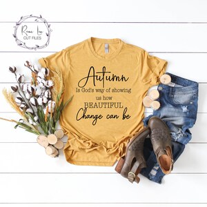 May include: A mustard yellow t-shirt with the text "Autumn is God's way of showing us how BEAUTIFUL Change can be" printed in black. The shirt is laid out on a white wooden surface with a bouquet of cotton and wheat, two wooden rounds, and a pair of brown ankle boots and blue jeans.