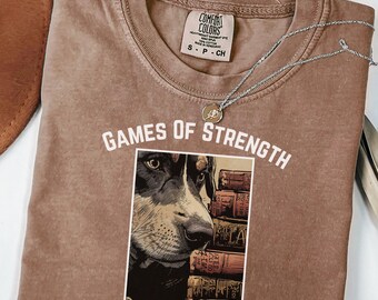May include: A brown Comfort Colors t-shirt with the text "Games of Strength Lawyer's Play." The shirt features a graphic of a dog's face with books in the background. A silver necklace with a pendant is draped across the shirt.