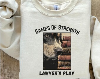 Peut inclure: Sweat-shirt crème avec le texte "GAMES OF STRENGTH" au-dessus d'un dessin de chien et de livres, et le texte "LAWYER'S PLAY" en dessous. Le sweat-shirt a un col rond et des manches longues.