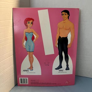 May include: A paper doll set featuring Ariel and Eric from the Disney movie The Little Mermaid. The dolls are printed on a pink background and have cut-out lines for easy assembly.