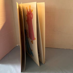May include: A book with paper doll cutouts of three dresses. The dresses are pink, blue, and white with a floral pattern. The book is closed and standing upright on a light blue surface.