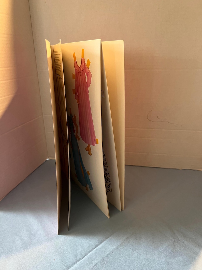 May include: A book with paper doll cutouts of three dresses. The dresses are pink, blue, and white with a floral pattern. The book is closed and standing upright on a light blue surface.