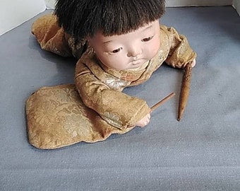 May include: A vintage porcelain doll dressed in a gold and brown patterned kimono, with black hair and a brown hat. The doll is holding a wooden stick.