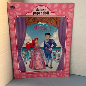 May include: A pink and white paper doll book featuring Disney's The Little Mermaid. The cover shows Ariel and Prince Eric standing on a balcony with a pink and white background. The book includes the text "Disney's The Little Mermaid deluxe paper doll".
