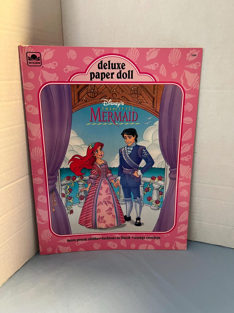 May include: A pink and white paper doll book featuring Disney's The Little Mermaid. The cover shows Ariel and Prince Eric standing on a balcony with a pink and white background. The book includes the text "Disney's The Little Mermaid deluxe paper doll".