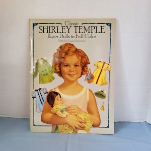 May include: Classic Shirley Temple Paper Dolls in Full Color book cover featuring a color illustration of Shirley Temple holding a doll. The book cover has a blue background with a white border.