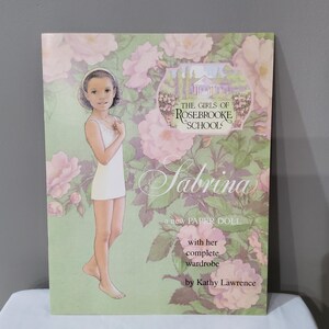 May include: A paper doll book cover featuring a girl named Sabrina, with a pink and green floral background. The text reads "The Girls of Rosebrooke Schools, Sabrina, a new paper doll with her complete wardrobe by Kathy Lawrence."