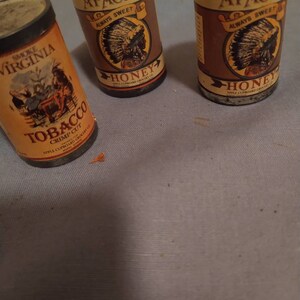 May include: Three vintage tobacco tins. Two tins are labeled "Apache Always Sweet Honey" and feature an illustration of an indigenous person wearing a headdress. The third tin is labeled "Smoke Virginia Tobacco Crimp Cut" and features an illustration of a horse-drawn carriage.