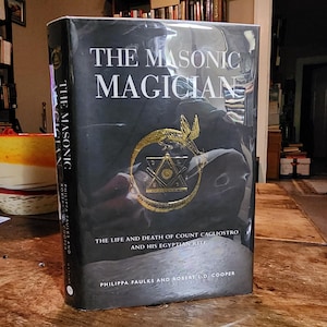 Puede incluir: Un libro negro de tapa dura titulado "The Masonic Magician: The Life and Death of Count Cagliostro and His Egyptian Rite" de Philippa Faulks y Robert L.D. Cooper. La portada presenta una serpiente dorada envuelta alrededor de un s&iacute;mbolo mas&oacute;nico.