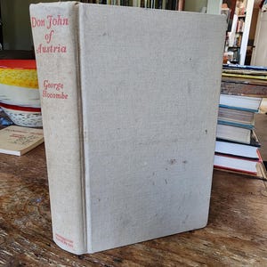 May include: A hardcover book titled "Don John of Austria" by George Slocombe. The book has a light beige linen cover with red lettering on the spine. The book is standing upright on a wooden surface.