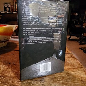 Puede incluir: La contraportada de un libro negro de tapa dura sobre la vida del conde Alessandro Cagliostro, una figura hist&oacute;rica conocida por su participaci&oacute;n en la masoner&iacute;a y la alquimia. El t&iacute;tulo del libro es "El Mago Mas&oacute;nico" y el texto describe la extraordinaria historia de Cagliostro, incluyendo sus viajes por Europa y su eventual condena a muerte por herej&iacute;a en Roma en 1789.