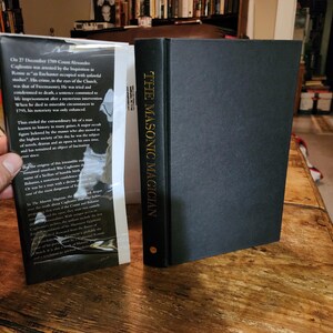 Puede incluir: Un libro negro de tapa dura titulado "The Masonic Magician" con letras doradas en el lomo. La contraportada presenta un texto sobre el tema del libro, Cagliostro, un ocultista italiano del siglo XVIII.