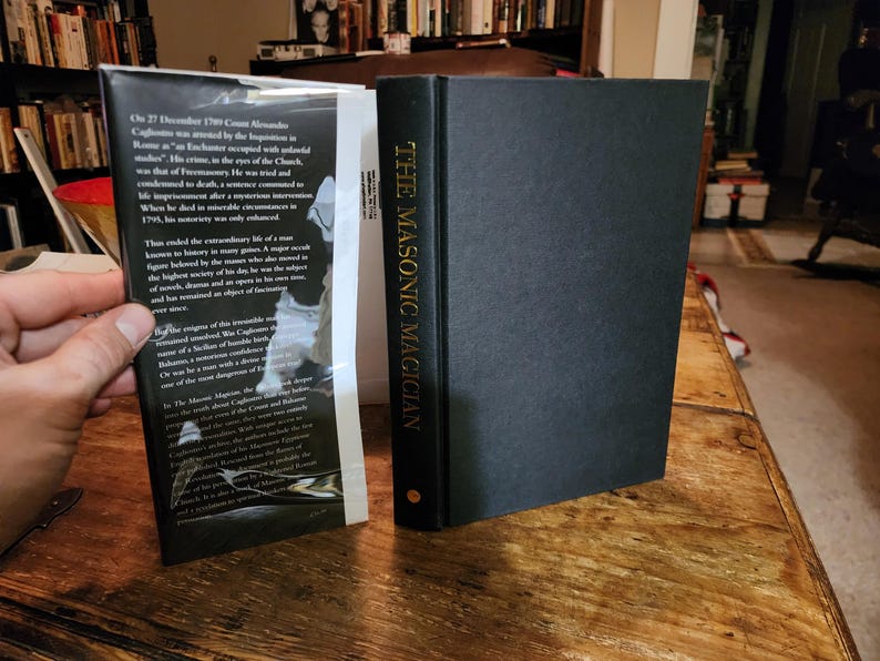Puede incluir: Un libro negro de tapa dura titulado "The Masonic Magician" con letras doradas en el lomo. La contraportada presenta un texto sobre el tema del libro, Cagliostro, un ocultista italiano del siglo XVIII.