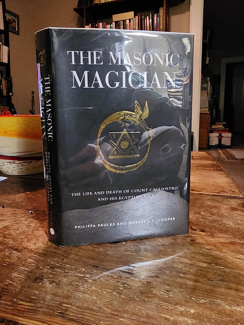 Puede incluir: Un libro negro de tapa dura titulado "The Masonic Magician: The Life and Death of Count Cagliostro and His Egyptian Rite" de Philippa Faulks y Robert L.D. Cooper. La portada presenta una serpiente dorada envuelta alrededor de un s&iacute;mbolo mas&oacute;nico.