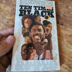Pode incluir: Um livro de bolso vintage intitulado "Ten Times Black" com uma capa com retratos de vários indivíduos negros. A capa do livro inclui texto sobre a experiência negra e a revolução cultural. O livro é segurado contra uma superfície de madeira.
