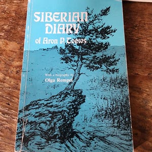 Pode incluir: Um livro de bolso intitulado Siberian Diary of Aron P. Toews com uma biografia de Olga Rempel. A capa apresenta uma ilustração a tinta preta de uma árvore solitária crescendo num penhasco rochoso sobre um fundo azul claro com texto branco.