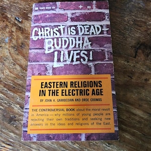 May include: A vintage paperback book with a brick-effect cover. The title "Christ is Dead Buddha Lives!" is in white lettering. An orange banner displays the title "Eastern Religions in the Electric Age". The book is by John H. Garabedian and Orde Coombs.