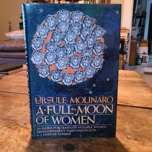 Peut inclure: Un livre bleu avec le titre "A Full Moon of Women" d'Ursula Molinaro. La couverture présente un motif circulaire de 29 visages féminins stylisés.