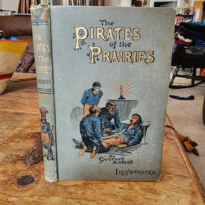 May include: An antique book titled "The Pirates of the Prairies" by Gustave Aimard. The cover is a muted teal colour with gold lettering and an illustration of men gathered around a candle. The spine shows a Native American illustration.
