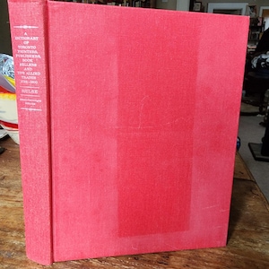 May include: A red hardcover book titled "A Dictionary of Toronto Printers, Publishers, Book Sellers and the Allied Trades 1798-1900" by Hulse. The book is standing upright on a wooden surface. The spine of the book is visible, displaying the title and author.