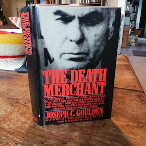 Puede incluir: Un libro negro de tapa dura titulado "The Death Merchant" con una imagen de primer plano de la cara de un hombre en la portada. El subtítulo del libro dice: "Intriga de la CIA, terrorismo internacional, acuerdos de armas multimillonarios - el mundo violento y sombrío que vio el ascenso y la caída de Edwin P. Wilson. Maestro traficante de armas ilícitas." El autor figura como "Joseph C. Goulden" con el texto adicional "Autor de The Superlawyers con Alexander W. Raffio".