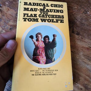 May include: A vintage paperback book titled "Radical Chic & Mau-Mauing the Flak Catchers" by Tom Wolfe. The cover is yellow with black text and a circular image of two people. The book's subtitle reads "Black Rage and White Guilt".