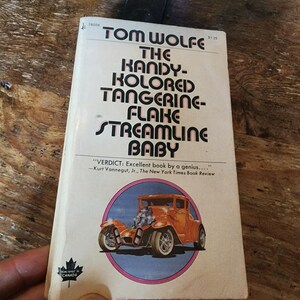 以下が含まれることがあります： トム・ウルフ著「The Kandy-Kolored Tangerine-Flake Streamline Baby」というタイトルのヴィンテージペーパーバック。表紙には、ピンクと青の円の中にオレンジ色のホットロッドカーのイラストが描かれています。