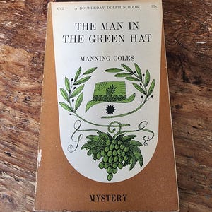 以下が含まれることがあります： マンニング・コールズ著「The Man in the Green Hat」というタイトルのヴィンテージペーパーバック。表紙には、緑の葉とブドウの房が付いた緑の帽子のイラストが描かれています。この本は「Doubleday Dolphin Book」で、下部に「Mystery」という言葉が印刷されています。