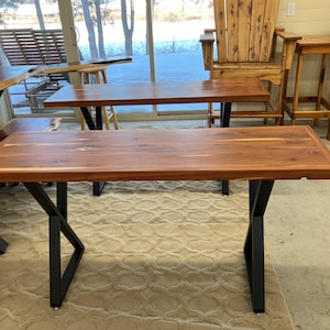 May include: A wooden table with a live edge top and black metal legs. The table is made of solid wood and has a natural finish. The legs are in a criss-cross design.