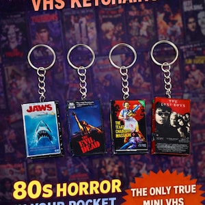 May include: Four mini VHS movie keychains featuring iconic 80s horror films: "Jaws," "The Evil Dead," "The Texas Chainsaw Massacre," and "The Lost Boys." Each keychain has a silver ring and chain. Text reads "80s Horror Movie VHS Keychains."