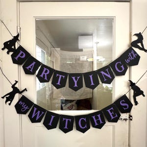 May include: Black banner with purple text that reads "Partying with my Witches." The banner is decorated with black witch silhouettes. The banner is hanging on a white door.