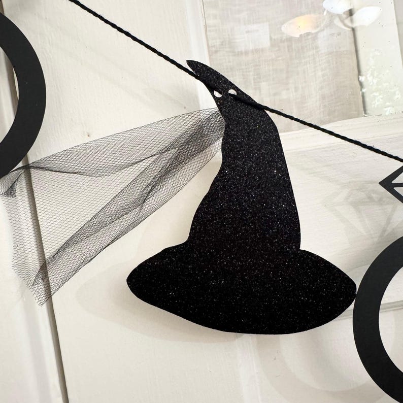 May include: Black glitter witch hat banner with a sheer black veil. The banner is strung on a black string and is perfect for Halloween decorations. The witch hat is a classic Halloween symbol.