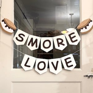 May include: A banner with the words "SMORE LOVE" in brown letters on white pennants. The banner is decorated with two s'mores, one on each side. The banner is hanging on a white door.