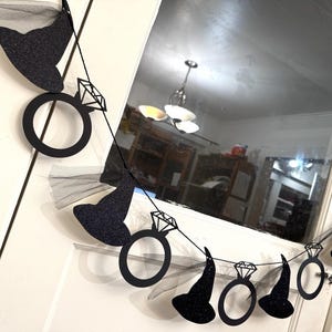 May include: A black glitter banner with witch hats and diamond rings. The banner is strung on a black string and is hanging in front of a white door and window.