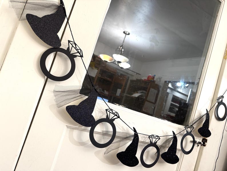 May include: A black glitter banner with witch hats and diamond rings. The banner is strung on a black string and is hanging in front of a white door and window.