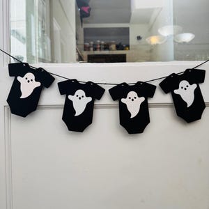 May include: A black Halloween banner featuring four onesie-shaped cutouts. Each cutout has a white ghost design. The banner is strung on a black string and is hanging in front of a window.