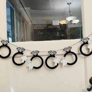 May include: A black Halloween banner with diamond ring shapes and white ghost cutouts. The banner is strung across a white door. The diamond rings are black with a geometric design. The ghosts are white with simple facial features.