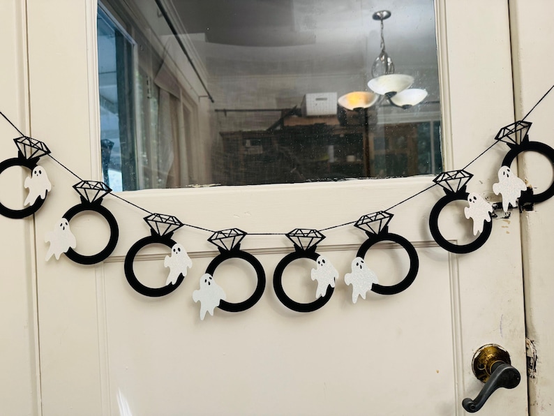 May include: A black Halloween banner with diamond ring shapes and white ghost cutouts. The banner is strung across a white door. The diamond rings are black with a geometric design. The ghosts are white with simple facial features.