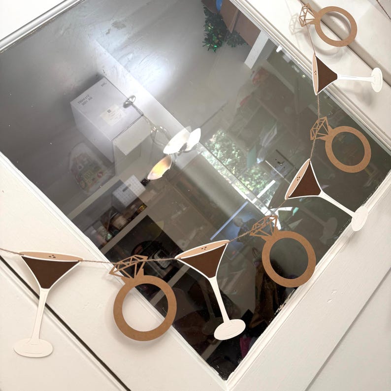 May include: A garland featuring cut-out shapes of martini glasses and diamond rings. The martini glasses have brown tops and white stems. The diamond rings are brown. The garland is strung on a thin string, perfect for a bridal shower or engagement party.