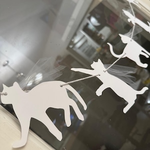 May include: A white cat-shaped garland with a string and tulle wings. The garland features multiple cat silhouettes, each with a small hole for stringing. The cats are connected by a thin, off-white string, and some have delicate tulle wings.