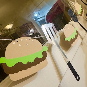 May include: A paper banner with a string of brown paper hamburgers, green lettuce, and brown patties. A silver spatula with a black handle is included in the banner.