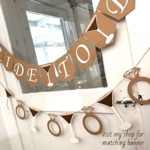 May include: A brown paper banner with white text that reads "Bride To Be". The banner also features martini glasses and diamond rings. The banner is hanging in front of a window.