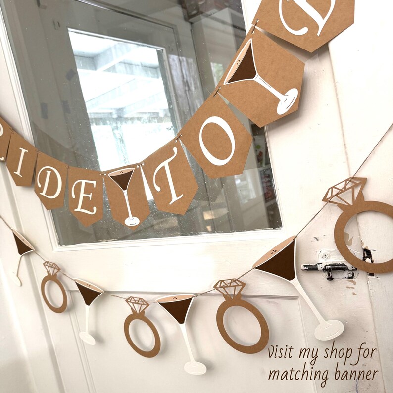 May include: A brown paper banner with white text that reads "Bride To Be". The banner also features martini glasses and diamond rings. The banner is hanging in front of a window.