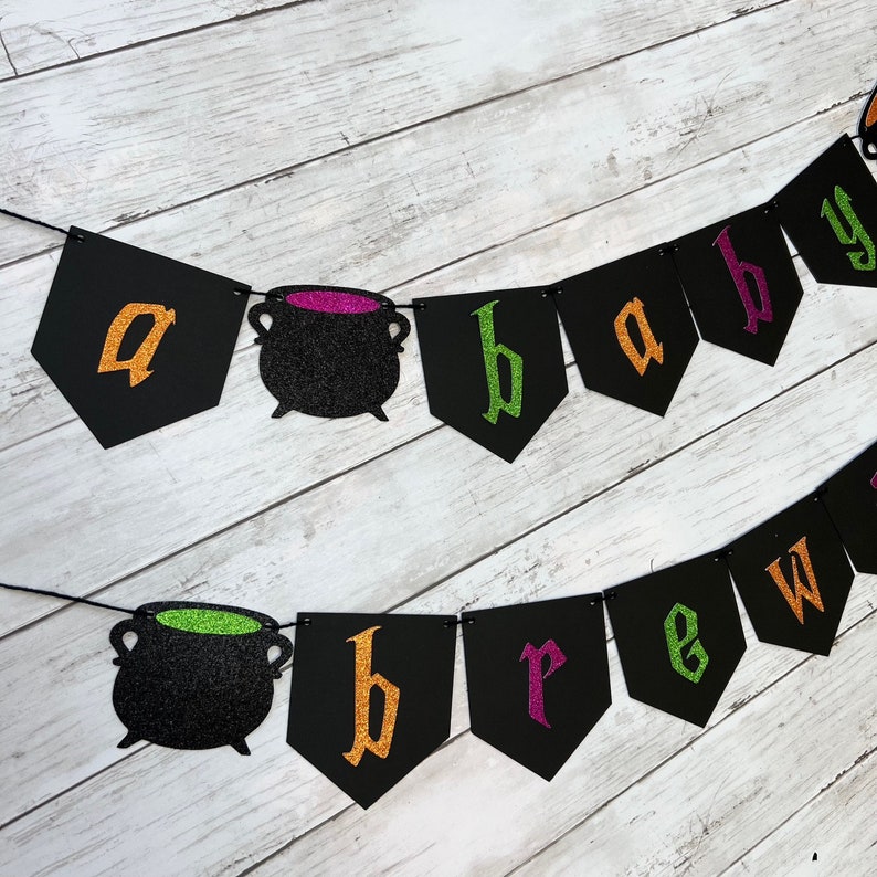 Puede incluir: Una pancarta de baby shower de Halloween negra y verde con el texto "a baby brew" y dos calderos negros con l&iacute;quido verde y morado.