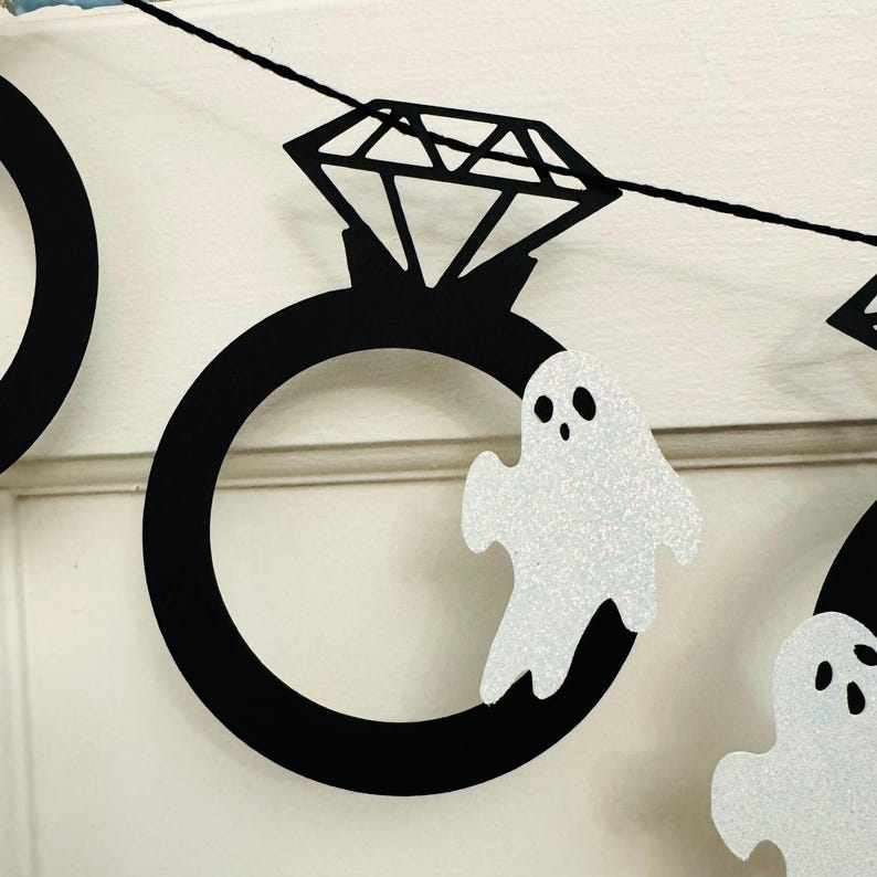 May include: A black and white Halloween banner featuring diamond ring and ghost cutouts. The diamond rings have a large black band with a diamond-shaped top. The ghosts are white with glitter and black eyes.