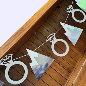May include: A silver glitter banner with diamond ring and triangle shapes strung on a white string. The banner is displayed against a wooden background, suggesting a celebratory occasion like a bridal shower or engagement party.