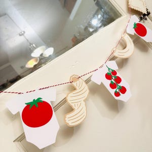 May include: A decorative garland featuring white baby onesies with red tomatoes and green leaves, and pasta shapes. The garland is strung on a red and white twine, perfect for a baby shower or Italian-themed party.