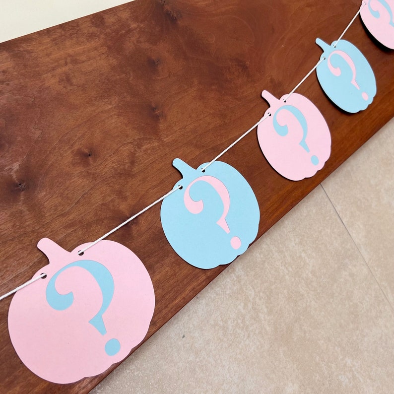 May include: A gender reveal banner with alternating blue and pink question marks in the shape of pumpkins. The banner is strung with white twine.
