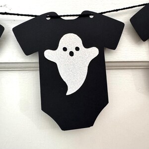 May include: Black Halloween-themed banner featuring a onesie shape with a glittery white ghost design. The banner is strung on a black string and is perfect for Halloween decorations. The ghost has three black dots for eyes and a mouth.