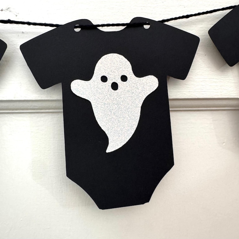 May include: Black Halloween-themed banner featuring a onesie shape with a glittery white ghost design. The banner is strung on a black string and is perfect for Halloween decorations. The ghost has three black dots for eyes and a mouth.